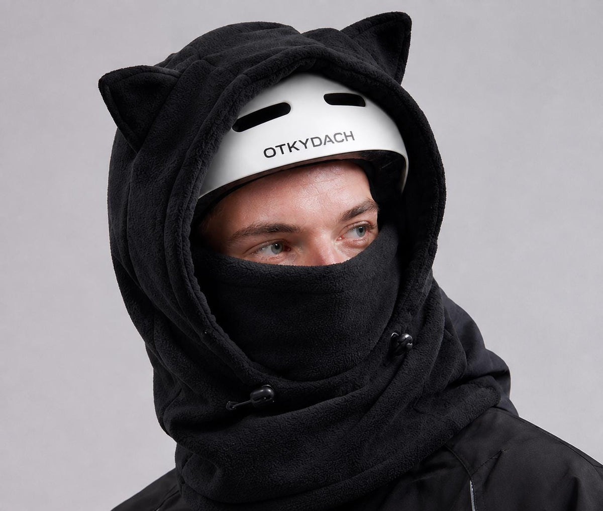 Black cat ear fleece hood with integrated face covering worn over ski helmet, adjustable drawstrings