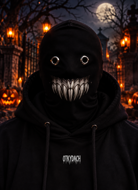 A person wearing a black hooded mask with eye holes and a wide row of sharp white teeth, photographed outdoors at night in a Halloween-lit cemetery. The hoodie has the word 'OTKYDACH' printed in white on the chest.